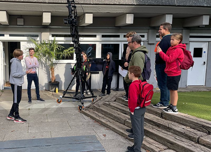 A group of people, including children and adults, are gathered outdoors in front of a building. Some kids have backpacks. A large camera crane is set up on the pavement. Everyone appears to be engaged in conversation or listening intently.