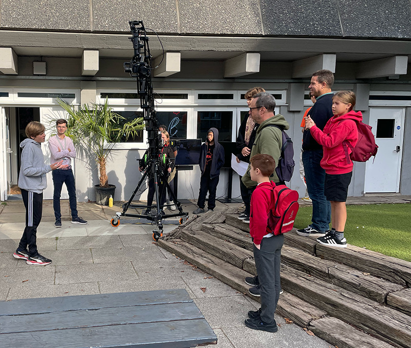 A group of people, including children and adults, are gathered outdoors in front of a building. Some kids have backpacks. A large camera crane is set up on the pavement. Everyone appears to be engaged in conversation or listening intently.