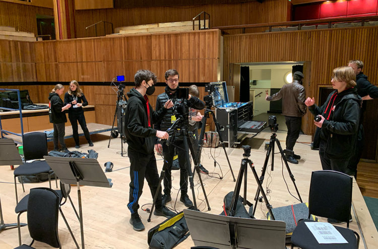 A group of students are setting up video and audio equipment in a multipurpose room with wooden walls. Some are handling tripods and cameras, while others adjust microphones. Chairs and music stands are scattered around, indicating a performance setup.