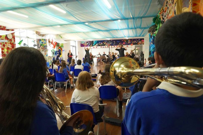 Students in a colorful, decorated classroom face forward while a conductor leads an orchestra. Some students in the foreground are holding brass instruments. Blue ceiling drapes and vibrant artwork add to the lively, creative atmosphere.