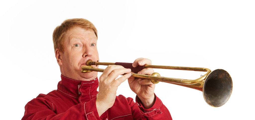 David Blackadder stands to the left against a white background. He wears a red jacket and is raising a trumpet to his mouth as he plays it.