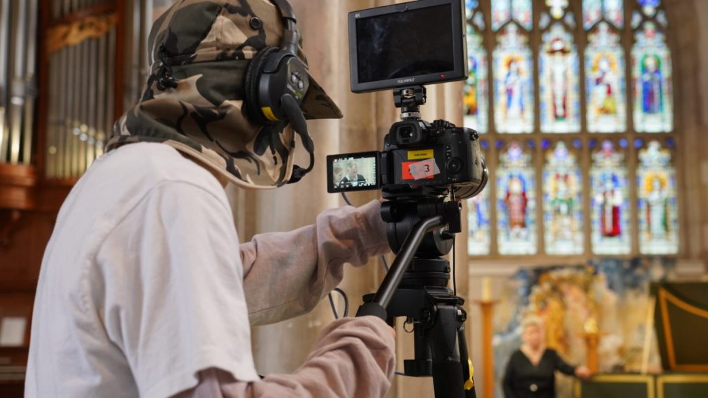 A student wearing camo headgear and a set of headphones operates a camera mounted on a tripod. The camera is directed towards a musician in the background standing in a church or cathedral with large, colourful stained glass windows.