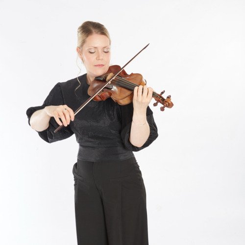 Julia Kuhn wears all black and stands before a white background. She holds her violin in position and looks down at its strings as she plays.