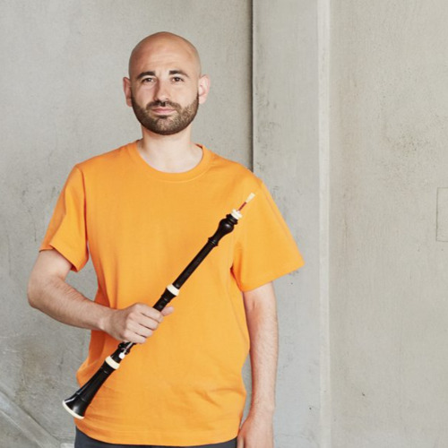 Leo Duarte wears an orange t-shirt and stands in front of a white background. He holds an Oboe in his right hand.