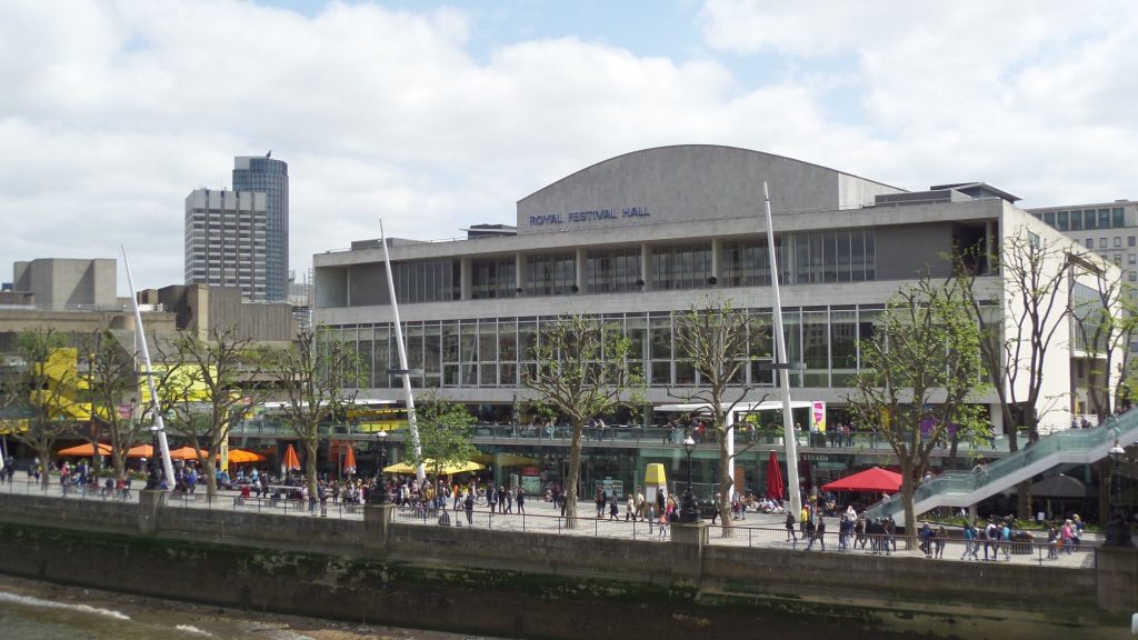 The image shows a view of the Royal Festival Hall in London from across the River Thames. People are gathered along the riverside, where outdoor seating and trees provide a vibrant atmosphere under a partly cloudy sky. High-rise buildings are visible in the background.