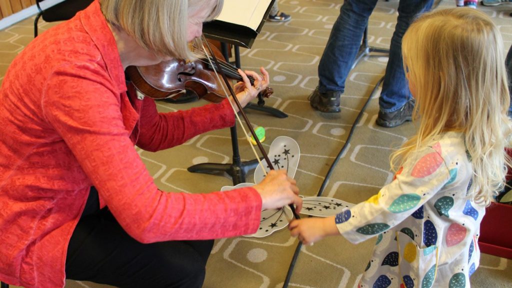 A woman with blonde hair, wearing a red jacket, is playing the violin while a young girl with long blonde hair, wearing a colourful dress, holds out a butterfly shape towards the violin. Both are in a room with a patterned carpet and other people in the background.