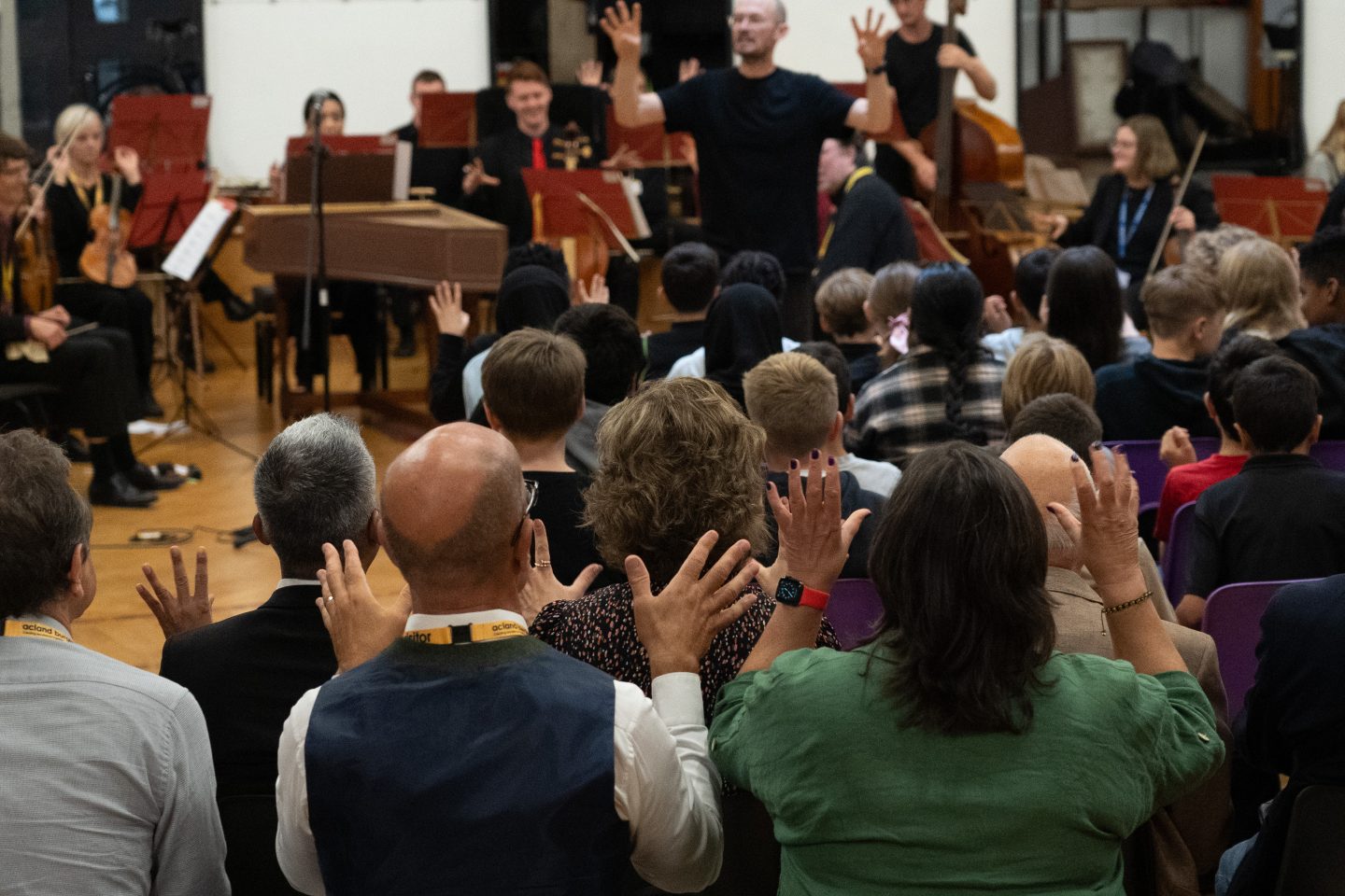 A diverse group of musicians and singers gather in a spacious, wood-paneled room. Some play instruments like drums and keyboards, while others are seated and standing. The walls are adorned with colourful artwork and posters.