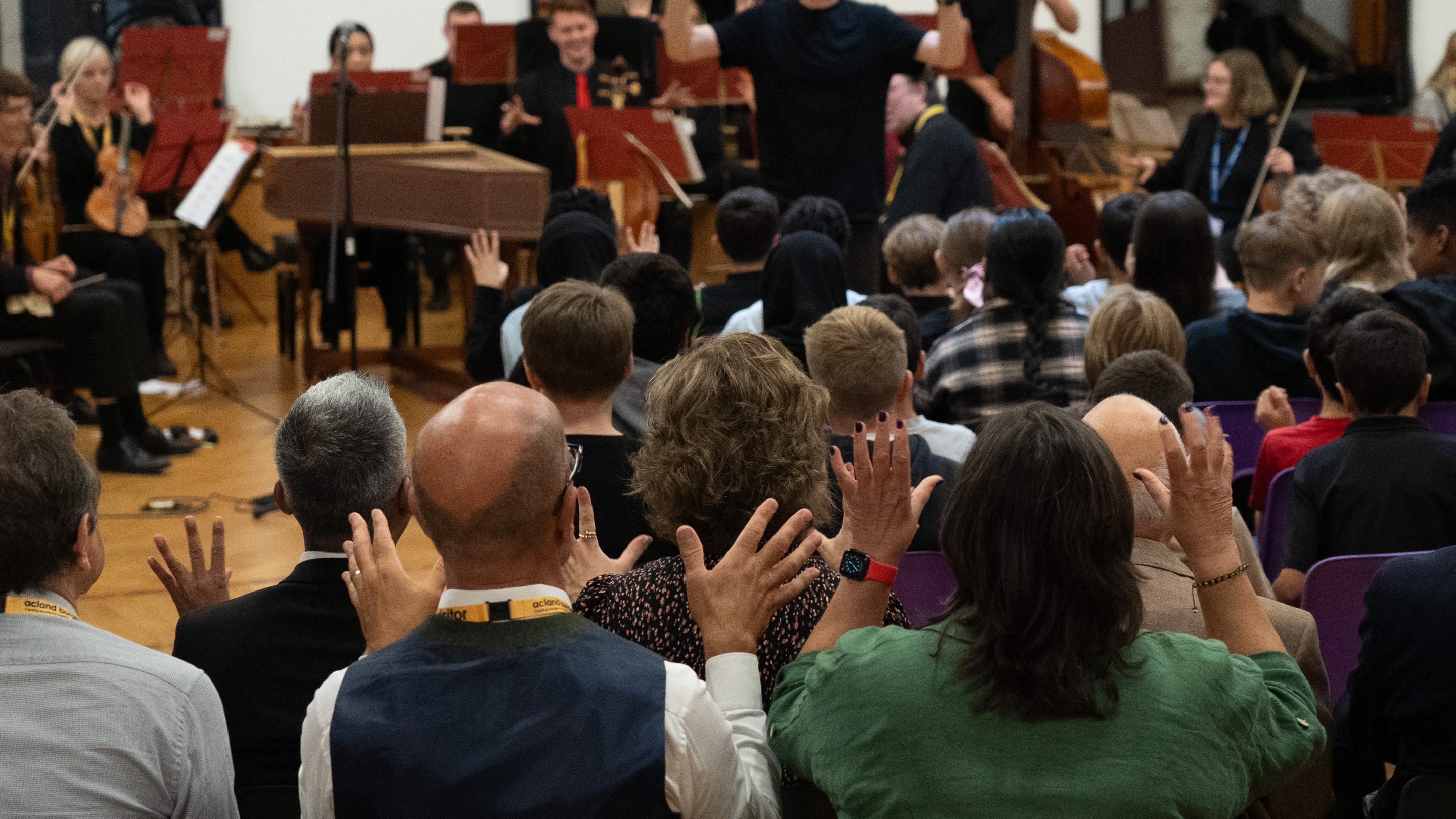 A diverse group of musicians and singers gather in a spacious, wood-paneled room. Some play instruments like drums and keyboards, while others are seated and standing. The walls are adorned with colourful artwork and posters.