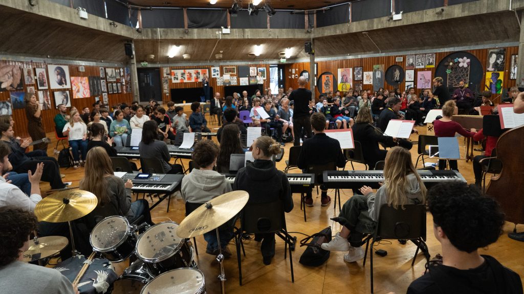 A diverse group of musicians and singers gather in a spacious, wood-paneled room. Some play instruments like drums and keyboards, while others are seated and standing. The walls are adorned with colourful artwork and posters.