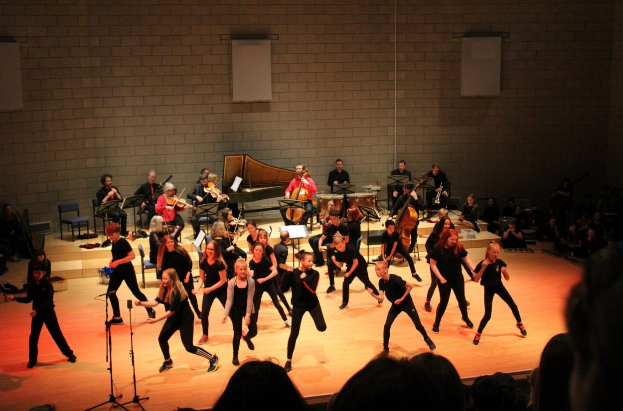 A group of performers on stage, dressed in black, energetically dancing in unison. Behind them, an orchestra with various musicians plays instruments. The stage is illuminated with warm lighting, highlighting the performers and musicians.