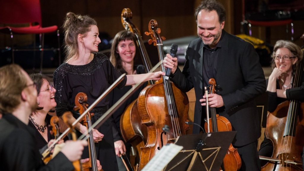 A group of smiling musicians in an orchestra, with a woman in black holding a microphone and passing it to a man. They are surrounded by cellists, and music stands with sheet music are visible.