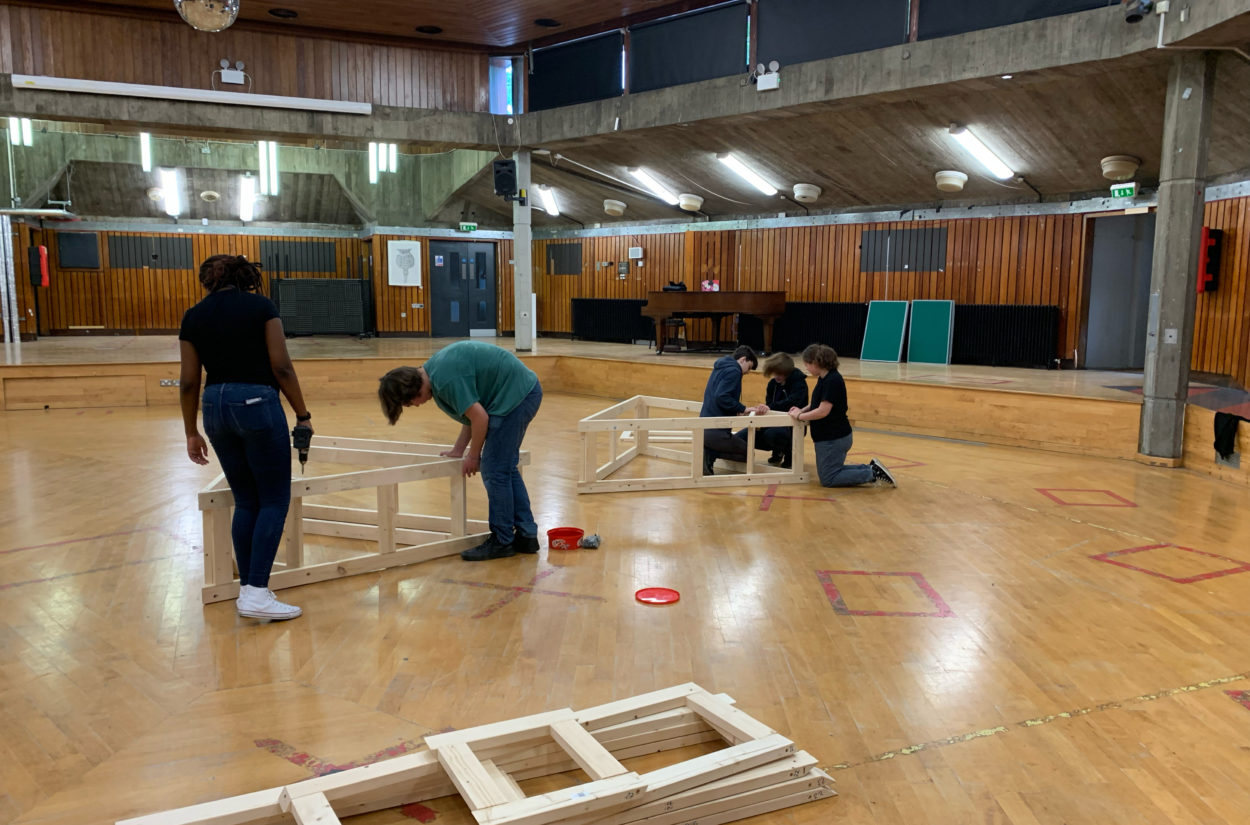 A group of people in a gymnasium are assembling wooden frames on the floor. The room has wood-panelled walls and bright overhead lights.