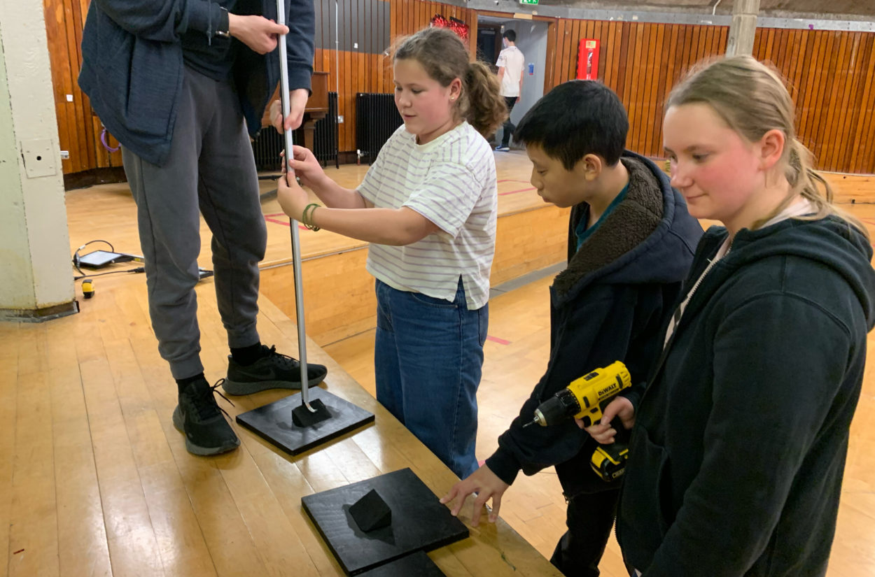 Four young people are assembling a stand in a wooden-floored room. One person is holding a pole, another is securing it, and a third is holding a drill. A fourth person watches. They appear focused on their task.