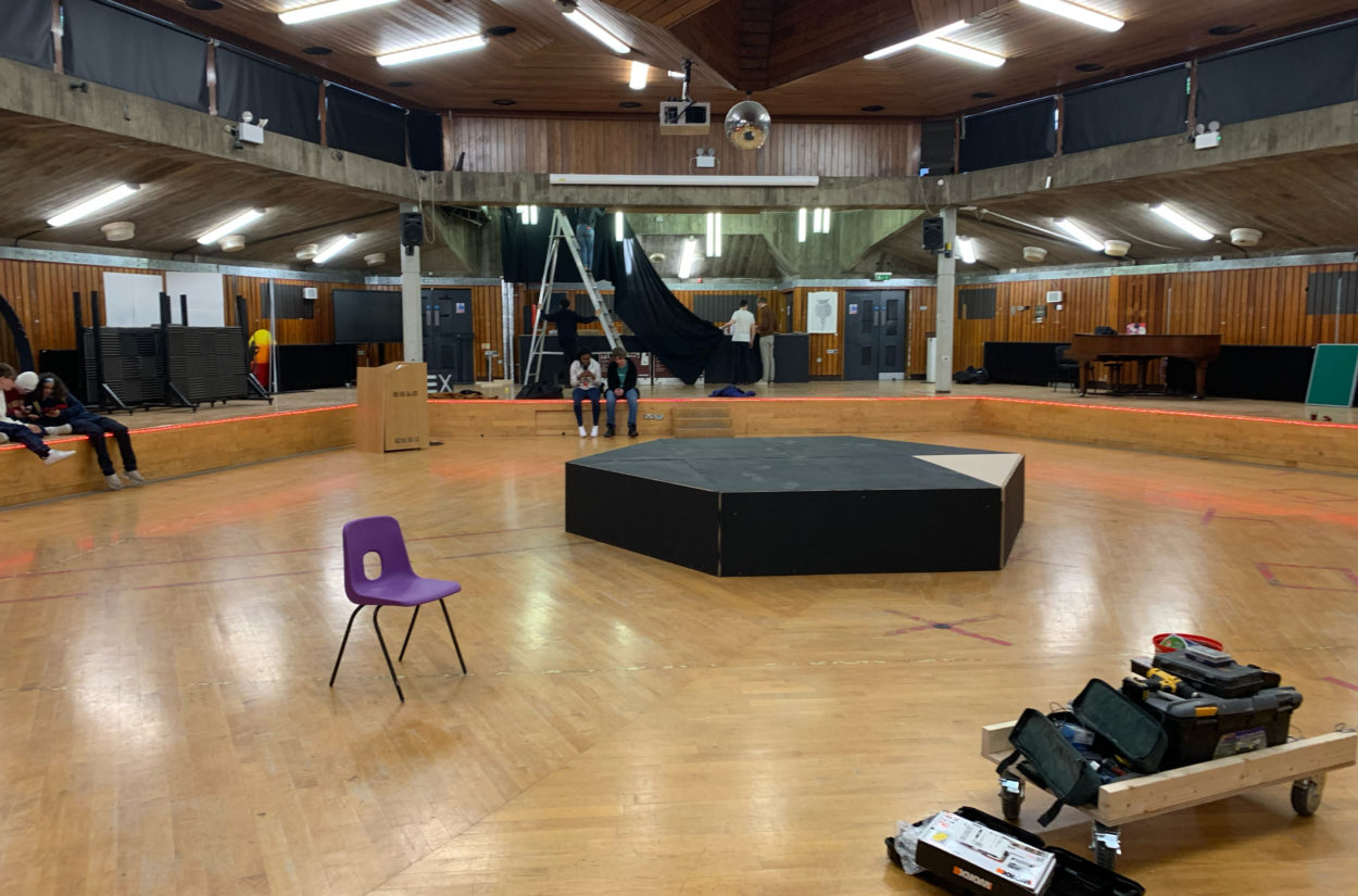 Interior of a circular hall with wood-panelled ceiling and floor. A hexagonal stage is at the centre. Chairs, tools, and equipment are scattered around. People are setting up the space.