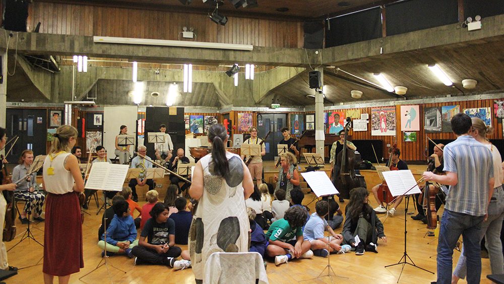 A group of musicians play instruments in a spacious room with wood walls and numerous seated children watching. Music stands and chairs are arranged throughout, and colourful artwork decorates the walls.