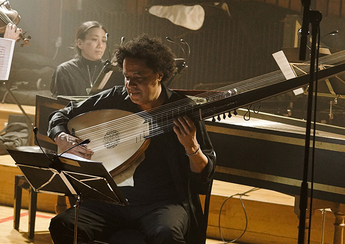 A musician plays a large lute in a dimly lit room, surrounded by music stands and sheet music. A harpsichord and another musician are in the background, creating an atmospheric setting for a classical performance.