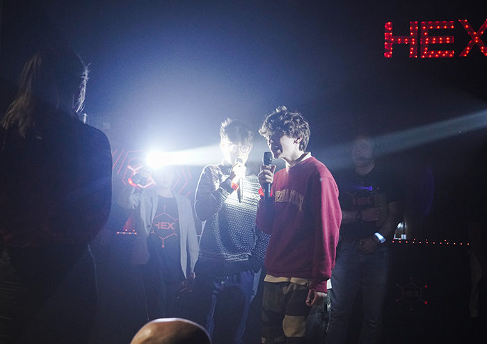 People stand in a dimly lit room with microphones, illuminated by bright lights. The background displays the word "HEX" in red.