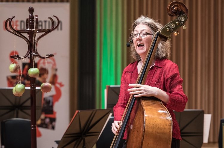 A woman plays a double bass on stage. She wears a red shirt and glasses, and appears to be passionately singing. A coat rack stands nearby, with apples hanging from it. Music stands and unoccupied chairs are in the background.