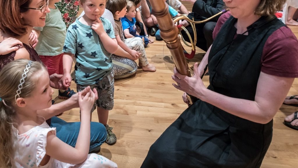 A woman plays a wooden wind instrument for a group of young children seated on the floor. A girl in a white dress touches the instrument, while other children watch in fascination. The setting appears to be an indoor educational or musical event.