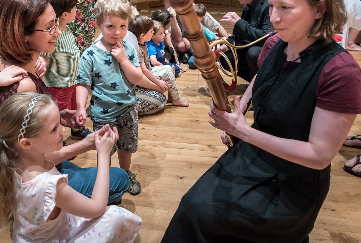 A woman plays a wooden wind instrument for a group of young children seated on the floor. A girl in a white dress touches the instrument, while other children watch in fascination. The setting appears to be an indoor educational or musical event.